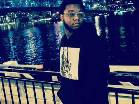 Lance Greenwood standing by the St. John's river at night, wearing a black hoodie near city lights.